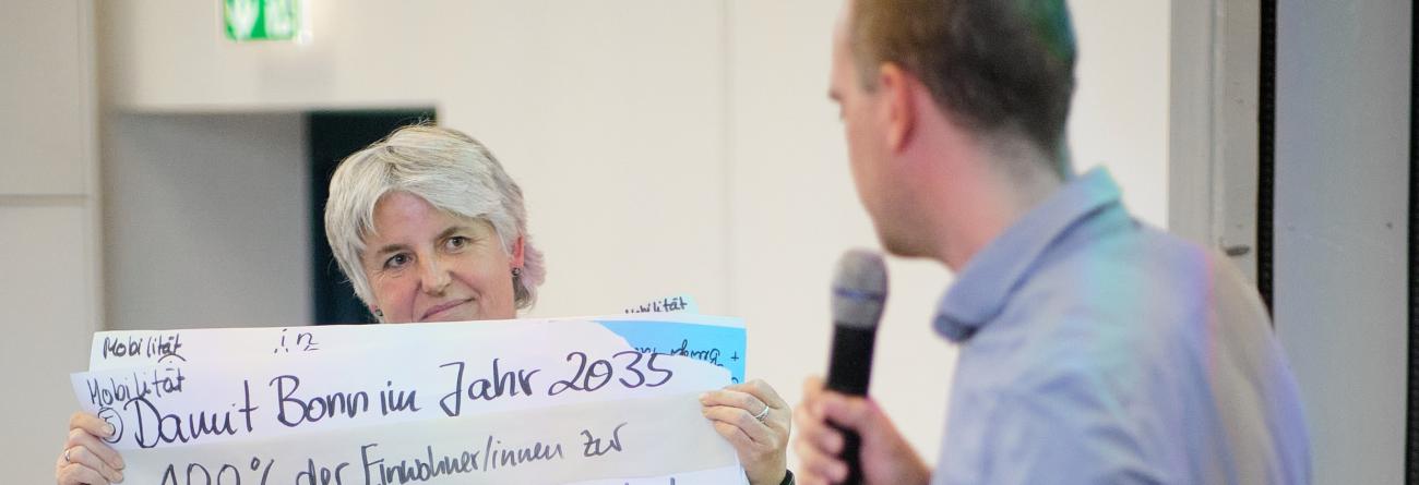 Abschlusspräsentation Eine Bürgerin hält ein großes Flipchart-Plakat hoch mit Forderungen für die Mobilitätswende, ein Mann mit Mikrophon liest es vor